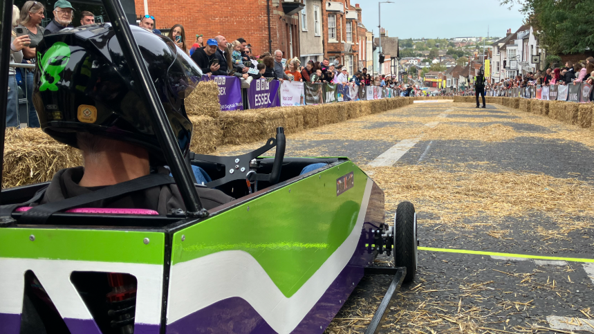 View standing behind a kart, that is on the left, which is sat at the top of a hill which has hay bales either side and people standing behind silver fences. The kart is painted green, white and blue.