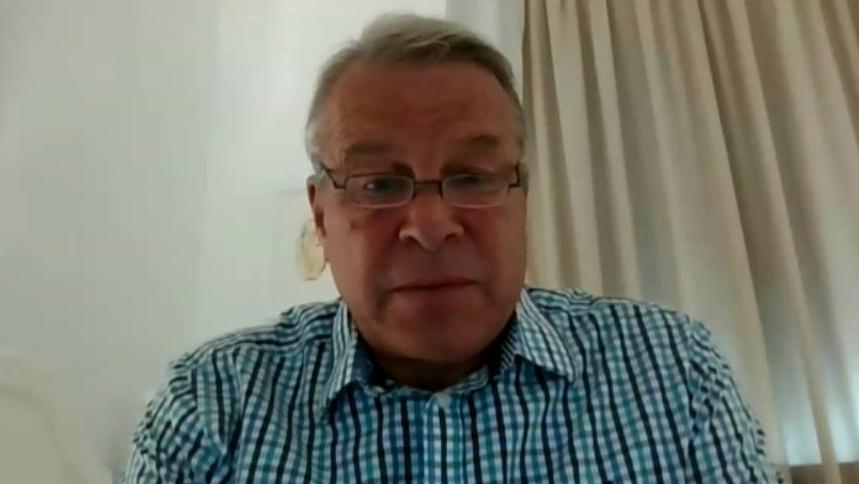 A Zoom screenshot of Chris Winter. He wears narrow glasses and a blue checked shirt, and is sitting in front of a pale wall with a light-coloured curtain drawn behind him. 