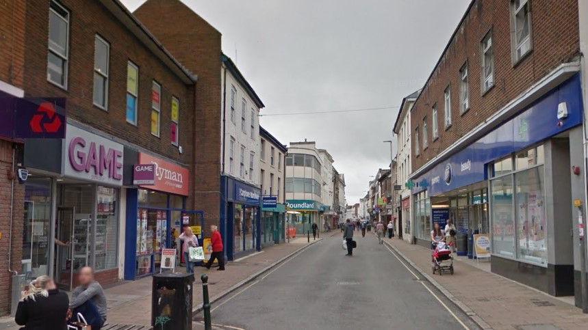 Google image of Barnstaple High Street. Retailers including Poundland and Boots can be seen along the street. People are walking along the pavement.