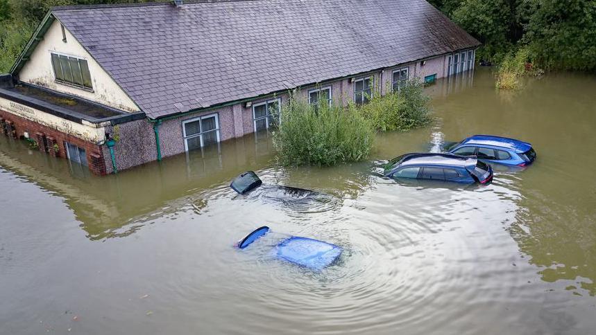 Drone footage of four cars submerged in water. There is a building in front where the water has reason to the first floor windows. 