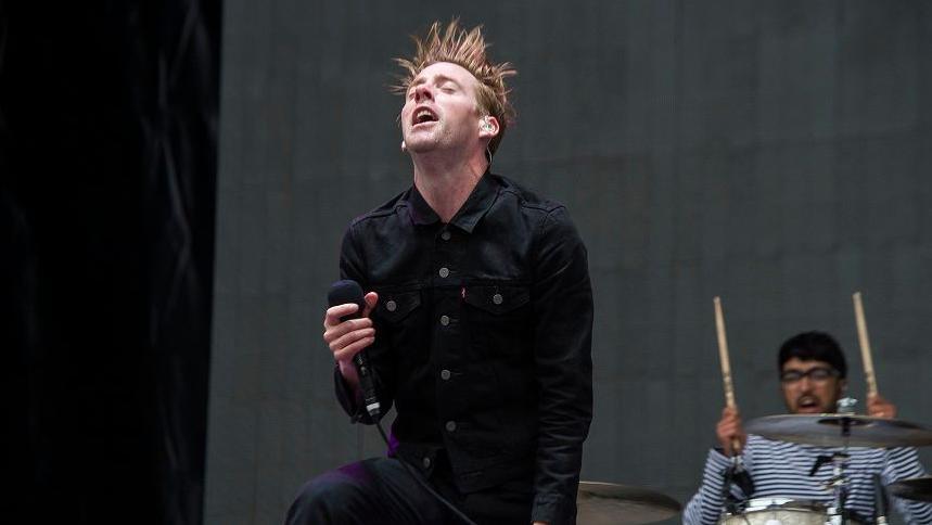 Ricky is performing mid jump, his hair is ginger and blowing in the air as he jumps. He is wearing a black shirt buttoned up and black trousers as he holds a microphone. Vijay is playing the drums wearing a blue and white lined long sleeved top. He has black short hair and is wearing glasses. 