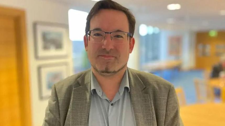 Lawrie Hooper, who has short brown hair and a thin goatee beard and is wearing glasses, a light coloured suit jacket and a grey open-necked shirt.