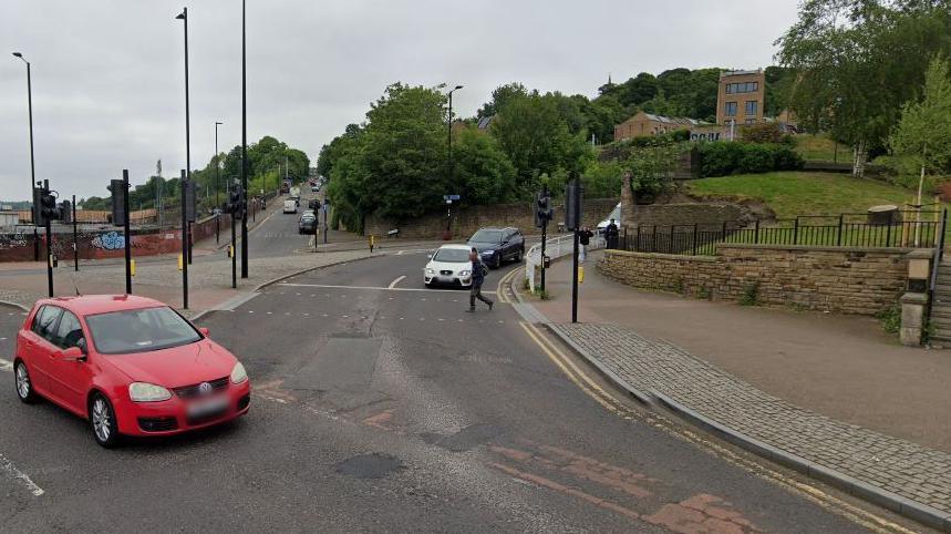 A road with another road joining on to it. A red car is on one road and a white and a black car are waiting at a junction on the other road. In the background there are trees and a large low-rise building.