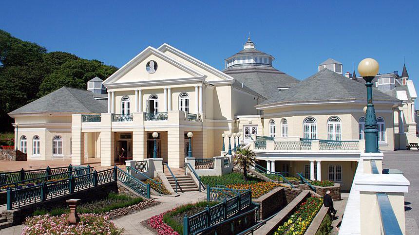 The Villa Marina, which is a large cream coloured building with grey tiled pointed roofing. There are stairs and walkways with metal railings in front of it that lead to flower beds and pathways.