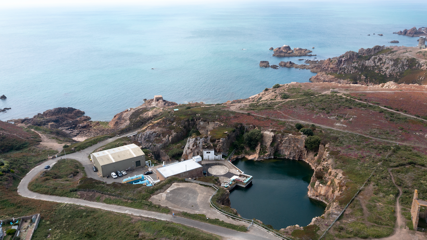 An aerial view of the desalination plant off the coast. A road can be seen winding down towards a large building with cars parked outside it and a body of water. Rocky surroundings face the ocean.