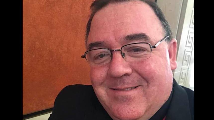 Fr Gallagher is smiling at the camera, in a selfie. He has short black hair and black glasses and is wearing a black shirt and robe.