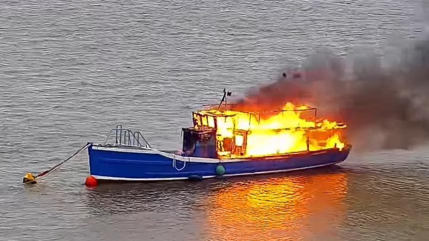 Large blaze on the Princess Marina ferry on the River Exe. The blue wooden ferry is moored up. Large orange flames tear through the wooden structure. Thick black smoke is pouring from the vessel.