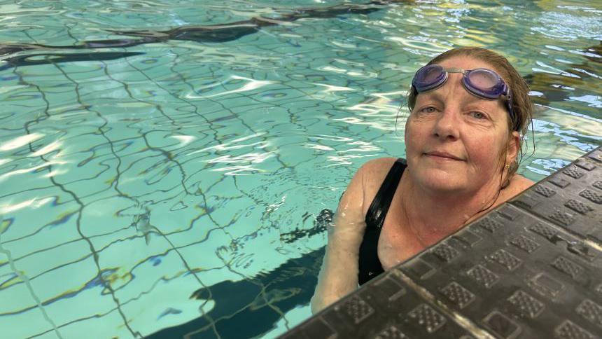 A woman with brown hair tied back, purple goggles on her forehead and a black swimming costume. She is in a pool and smiling into the camera from the edge. You can see part of the surface of the swimming pool in the background.