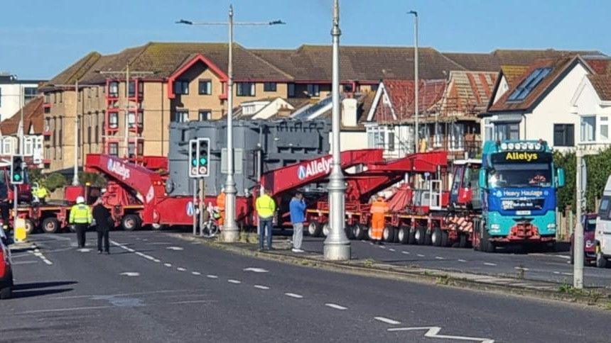 The convoy lorry is seen from across the street and is a very long lorry with grey industrial equipment in its middle part. There are people standing around the lorry in the road, some wearing hi-viz clothing.