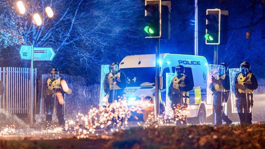 Five police offiers with helmets and riot shields in front of a police van at traffic lights. Sparks of a firework hit the road in front of them. This is a night time shot.
