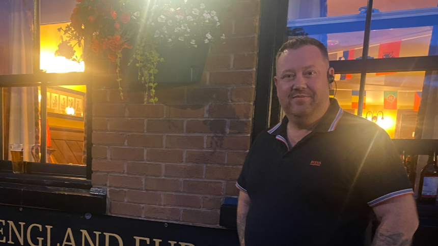 Steve, standing outside of his pub, there are two windows behind him, separated by a wall on which a flower basket is hanging