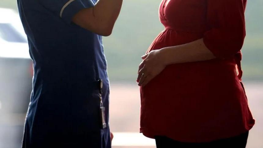 A nurse in a blue uniform stands and talks to a pregnant woman in a red top, who has her hand on the bump. Only the abdomens and arms can be seen