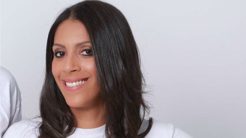 A woman with dark shoulder length hair stands in a white T-shirt. She has a broad smile.