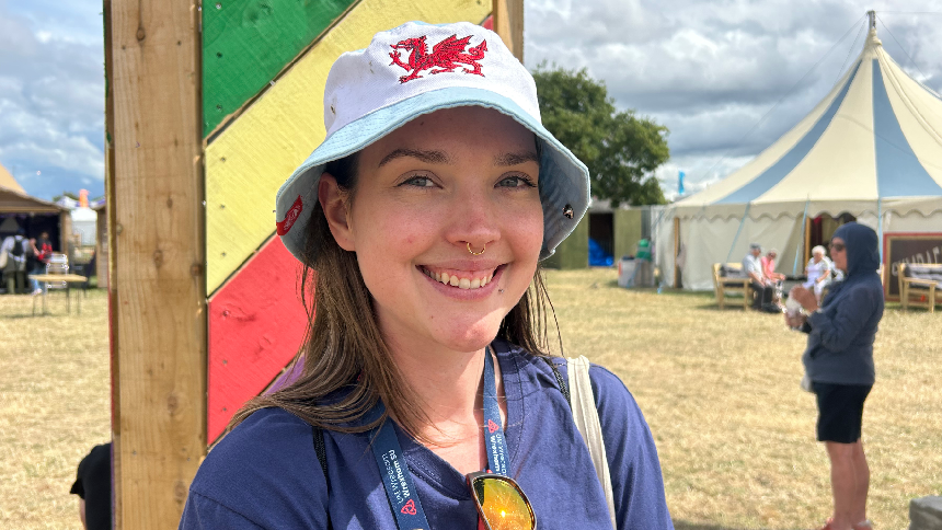 Close up image of Ariana smiling at the camera. She is wearing a blue t-shirt and a light blue bucket hat which has a red dragon on. Ariana has a Wrexham University lanyard around her neck. She is standing in front of the learners area at the Eisteddfod.