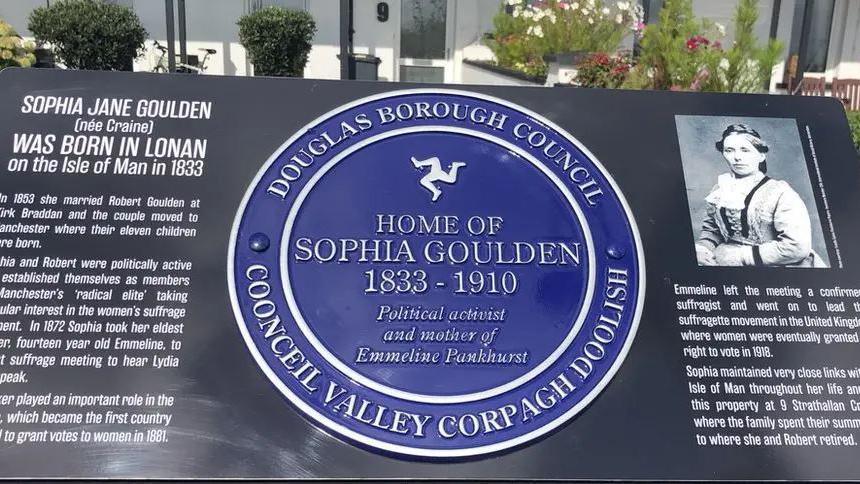 A blue circular plaque reading "Home of Sophia Goulden 1833-1910" with a Manx triskelian above it. To the left and right of the plaque is a large black sign with information about Mrs Goulden in white lettering. Behind the sign you can see a front door and white exterior walls with various plants.