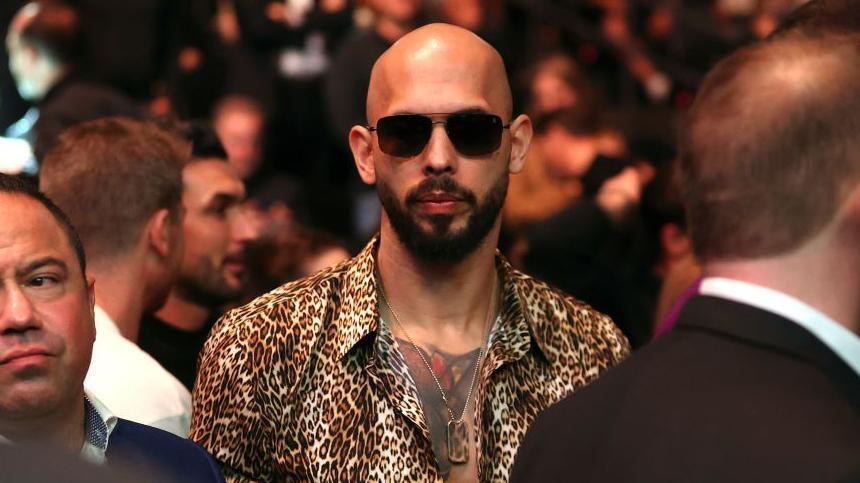 Andrew Tate wearing dark sunglasses and a button-down animal print shirt and a dogtag necklace