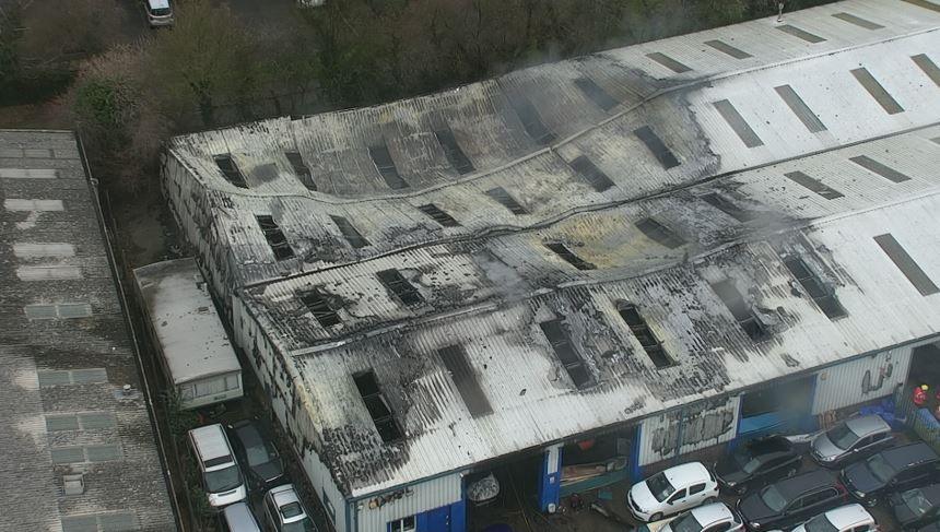 An aerial shot show a charred and partially collapsed metal roof on a large warehouse. There are cars and a lorry parked around the sides of the building