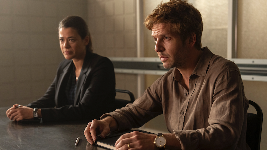 Damien Molony alongside co-star Celine Arden, sat side by side at a desk looking serious in a dark room.