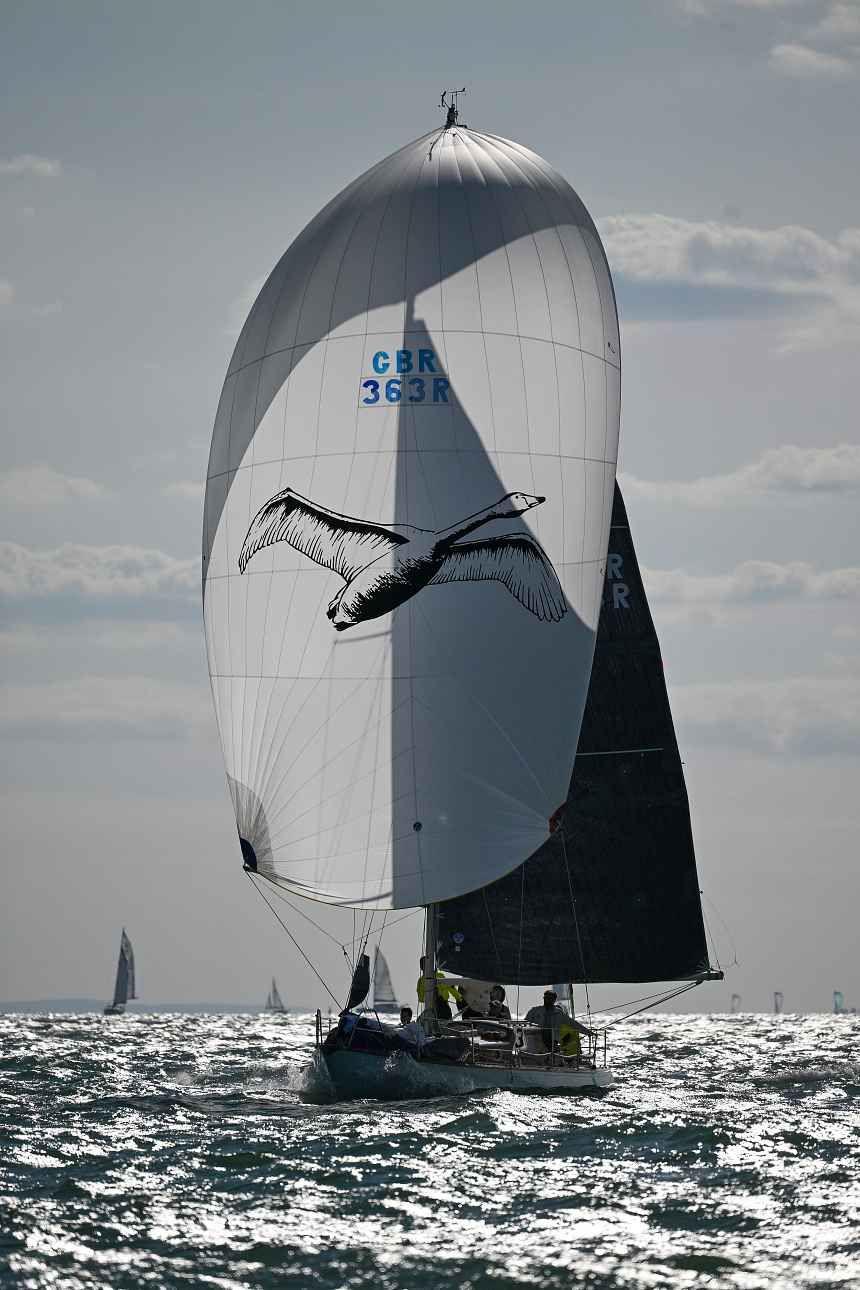 Racing yacht Whooper on the water during Cowes Week. It has a large white billowing sail bearing a picture of a flying swan.