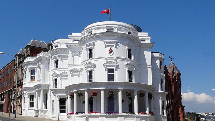 The exterior of the Tynwald legislative buildings, a grand round white structure with colums at the bottom, a Manx glag flies at the top, on a clear blue day.