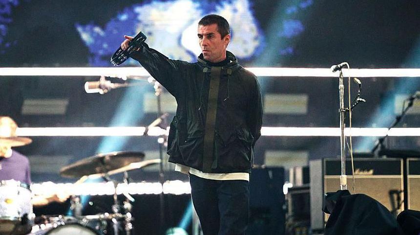 Liam Gallagher pointing with his right hand into the crowd at an Oasis concert. He is holding a tambourine in his right hadn and is wearning a dark coloured jacket.