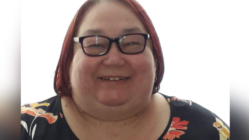 Wendy Gane smiling at the camera. She has red hair in a chin-length bob cut and wears black-rimmed glasses. She is wearing a black top with orange and yellow flowers on it.