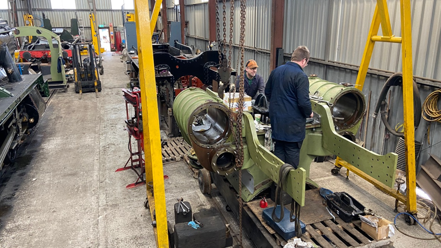 Inside a railway works shed with two people working on a large piece of locomotive machinery