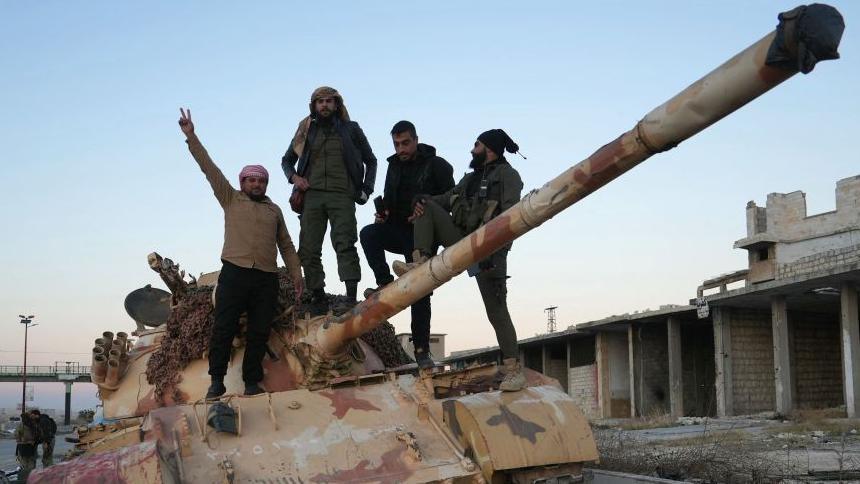 Four men stand on top of an abandoned tank on the road to Aleppo. One man on the left has his hand raised in a v-for-victory style salute.