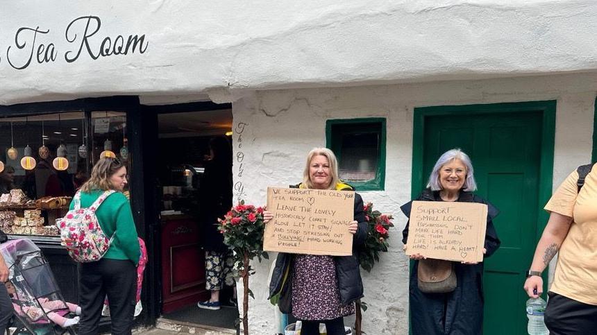 Two supporters hold up placards outside the tea room, reading "leave the lovely, historically-correct signage alone" 