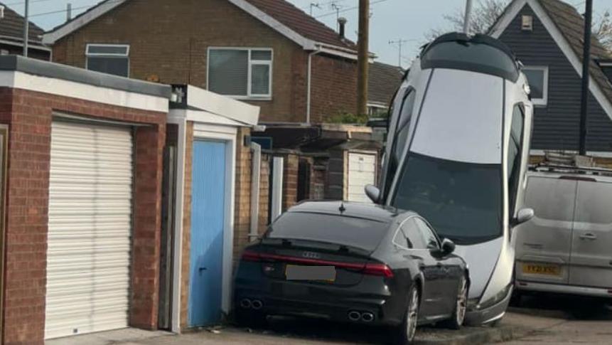 A black Audi car parked up against a silver car, which is impaled upright against a lamp-post. Both vehicles are are next to a row of a garages with the shutters closed and in the background are a line of houses with only the rooftops visible