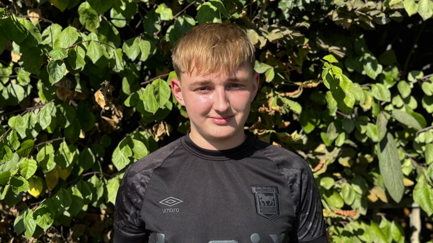 A head and shoulders image of Baily Wie. He is wearing a black Ipswich Town shirt and smiling into the camera. He is standing in front of trees.