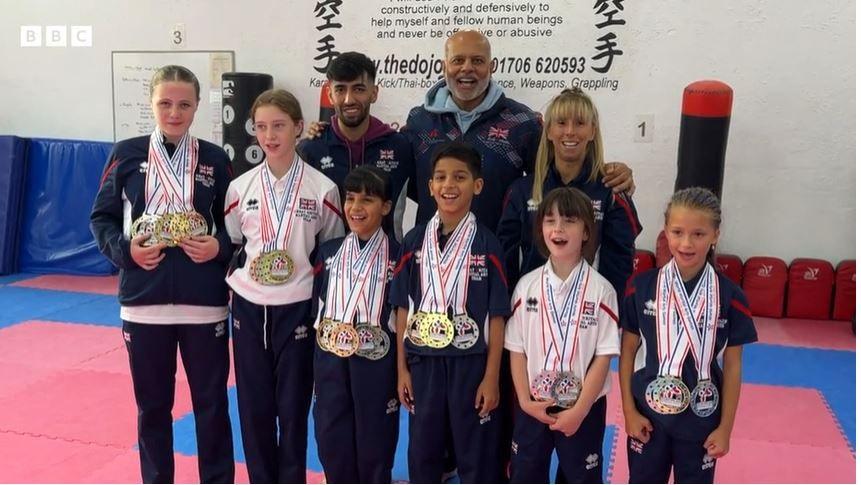 Six children stand in front with their karate medals while three teachers stand behind them smiling