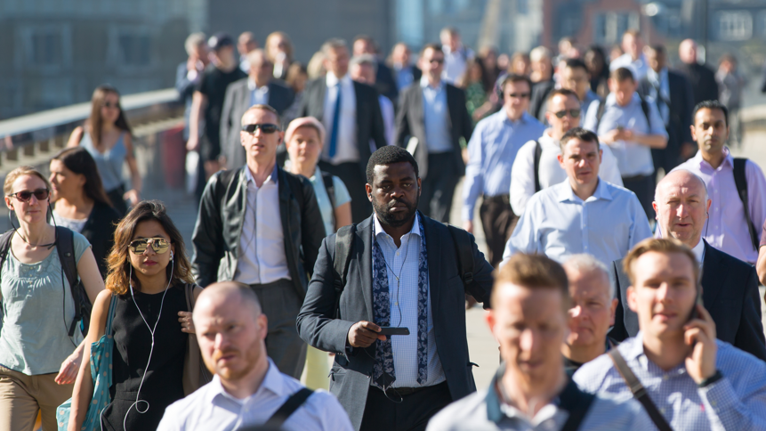 men and women mostly smartly dressed in shirts and jackets walking towards the camera seemingly in London over a bridge in the sunshine