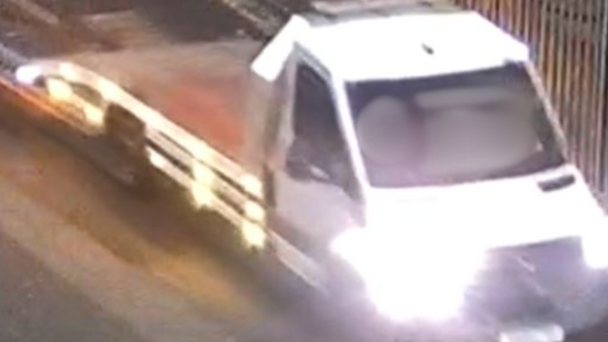 A blurred image of a van with a white cab, and an open flat back. The headlights are shining.
