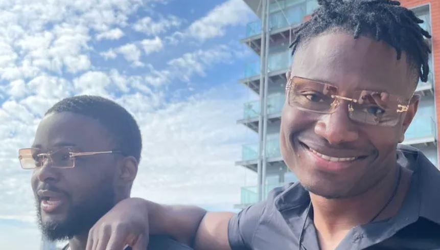 A close-up of two people wearing matching orange-tinted sunglasses. The man on the right is smiling and has his arm on the shoulder of the man on the left, who looks happy but is looking away from the camera. Clouds are in the blue sky and a block of flats is in the background.