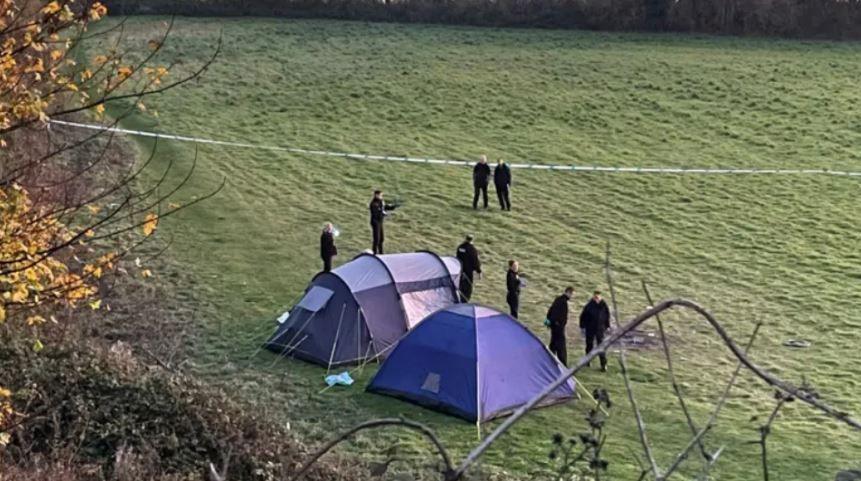 Two blue tents in a field are cordoned off by police tape. Eight people in dark clothes are standing close to the tents behind the police tape