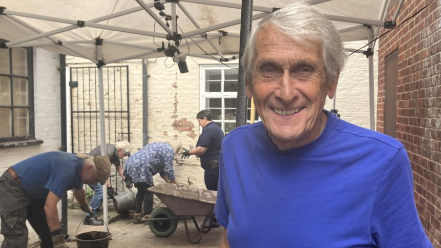 Archaeologist Peter Allen looking at camera while team digs behind him. There is a gazebo above and a wheelbarrow and buckets being used by the people in the background.