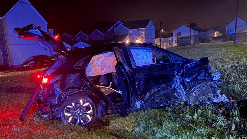 A badly damaged black vehicle in a patch of grass. The front of the car is completely torn off, the car door on the back right hand side is open, along with the boot door.