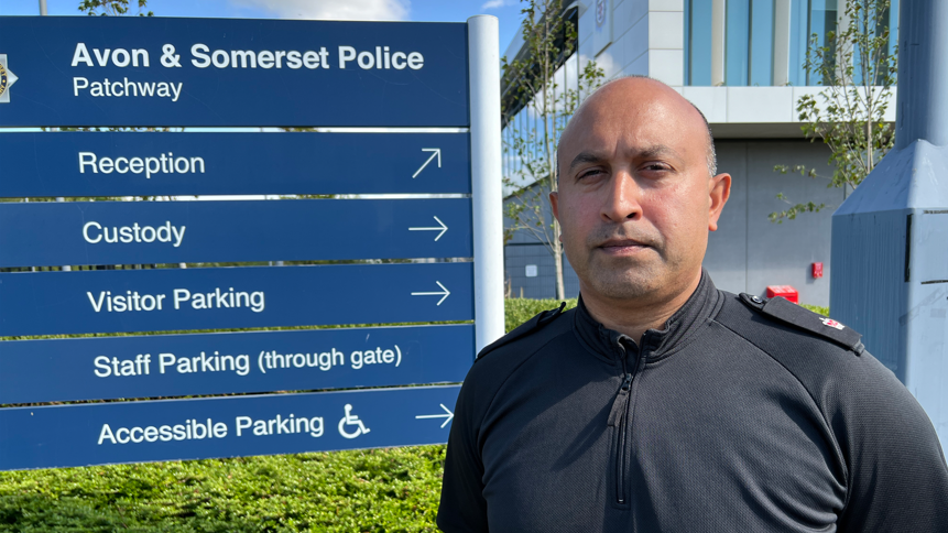 Supt Int Deepak Kenth is wearing a black shirt, stood outside the entrance to an Avon and Somerset Police office. 