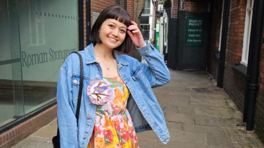 Nina , a young woman smiling at the camera. She is wearing a floral colourful dress with a denim jacket over it.