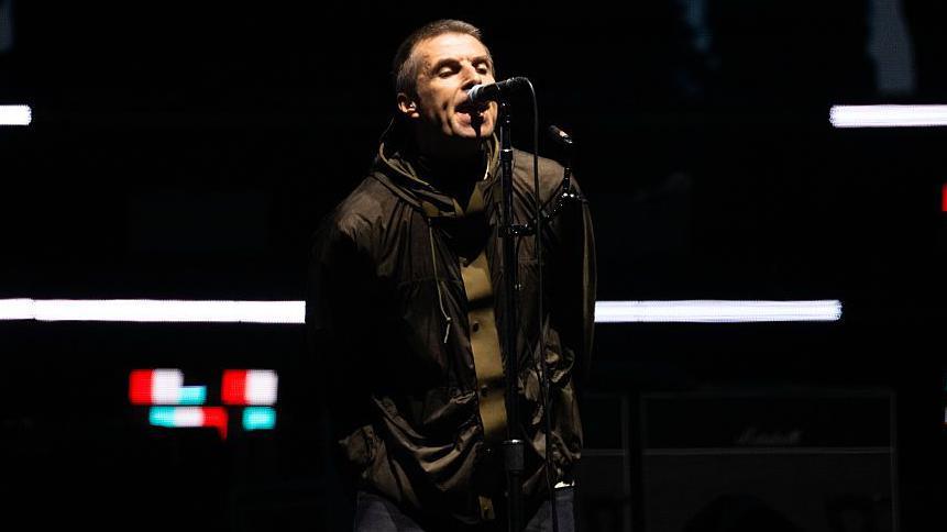 Oasis singer Liam Gallagher sings into a microphone with his hands behind his back at the opening night of the band's reunion tour in Cardiff, Wales