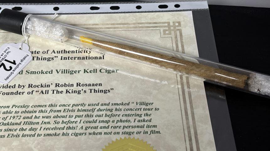 A long cigar with a yellow end concealed in a tube and resting on a certificate for the item.