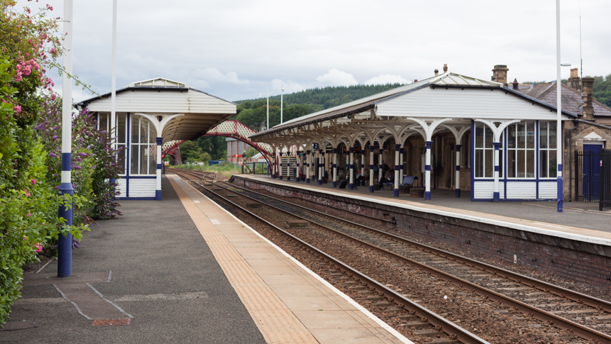 A picture of Hexham station with blue and white Victorian station buildings on either side and a footbridge 