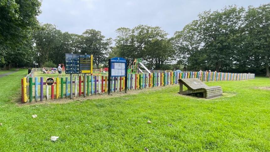 A multicoloured fence encloses a play area. In the distance you can see a slide and a climbing frame. It is in a grassy area.
