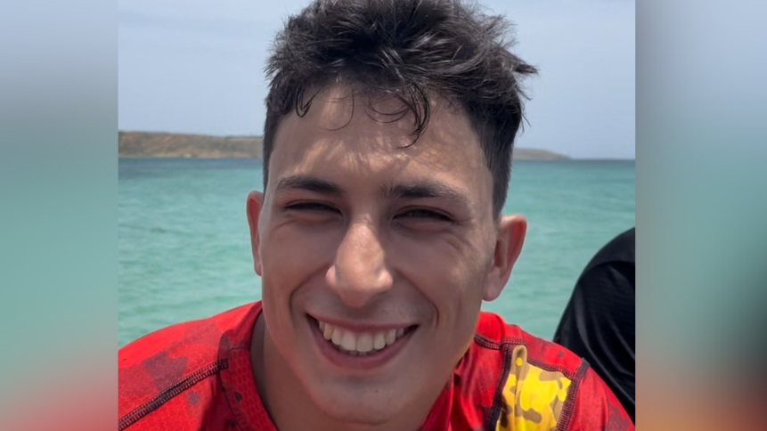 Sebastian Pena is young with short brown hair. He is wearing a red and yellow swimming top. Behind him is the sea and land is on the horizon.