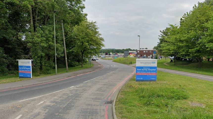 An external shot of East Surrey hospital with signs either side of the road and low brick buildings seen on the hospital site. There are grass verges and trees on either side of the road.