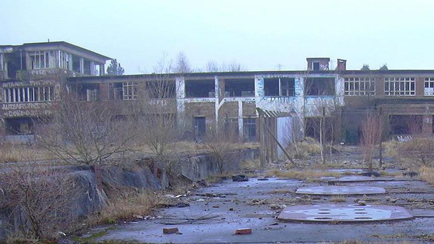 The deserted site of Huncoat power station. It is a long two-storey brick building with a taller section to the left which have been vandalised. To the front are overgrown flat areas with covers on the ground and sprouting weeds