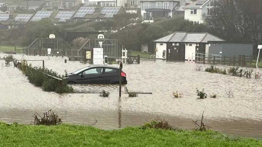 A black car parked in a flooded parking area. The water is covering the wheels. There is a park in the background and small buildings on the right