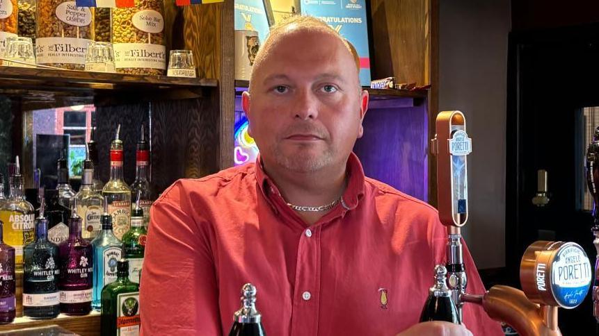There is a man in a pink polo shirt and a chain. He is stood behind a bar whilst holding a tap. He has very short white hair and light goatee facial hair. Behind him, to his left, is a number of spirits, including Absolute vodka and Jägermeister
