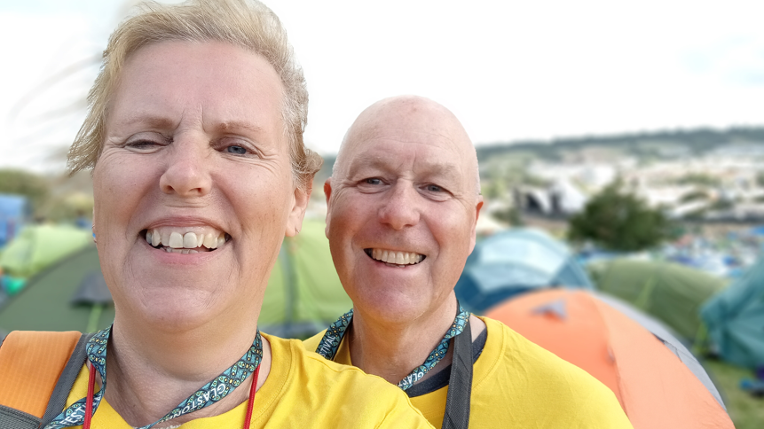 A woman taking a selfie, smiling at the camera, with her partner in the background. They are standing in a campsite among many tents at Glastonbury Festival.